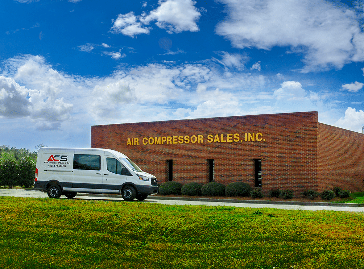 A white van parked outside a brick building with large yellow letters reading AIR COMPRESSOR SALES, INC. on a sunny day.
