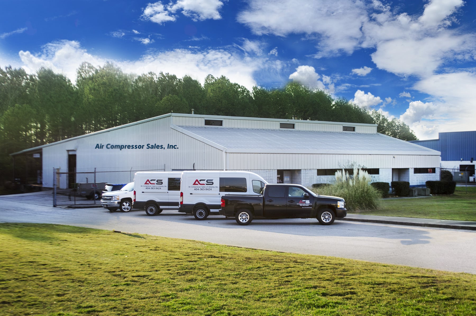 A large white industrial building labeled Air Compressor Sales, Inc. with three company vehicles parked outside on a sunny day.