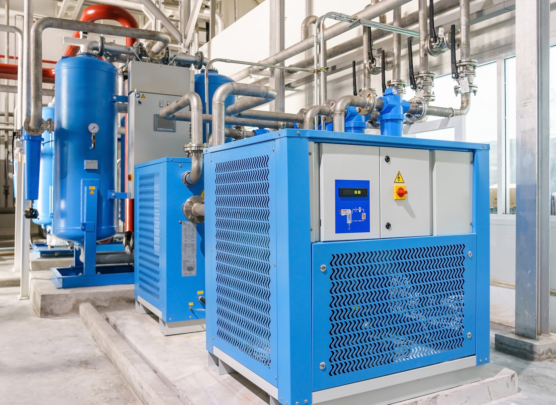 Blue air compressor units connected to metal pipes, indoors on a concrete floor, with control panels and visible pressure gauges.