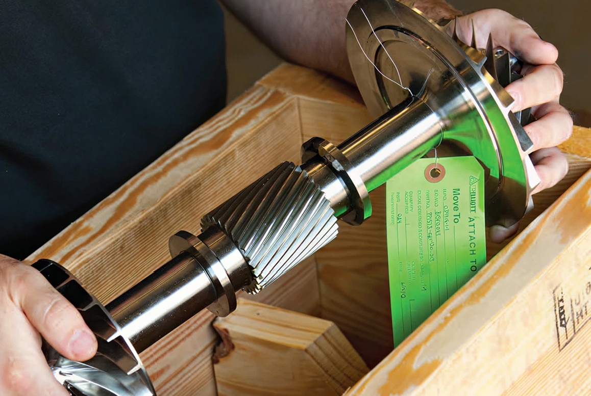 A person holds a large shiny metal gear shaft with precise teeth, partly in a wooden crate. A green inspection tag is attached.