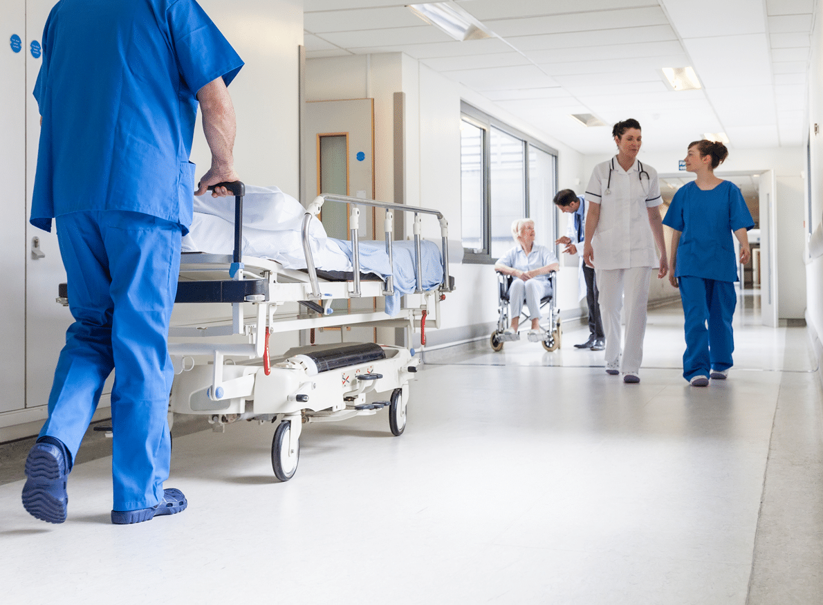 A healthcare worker pushes a gurney down a hospital hallway while two nurses and a patient in a wheelchair walk behind.