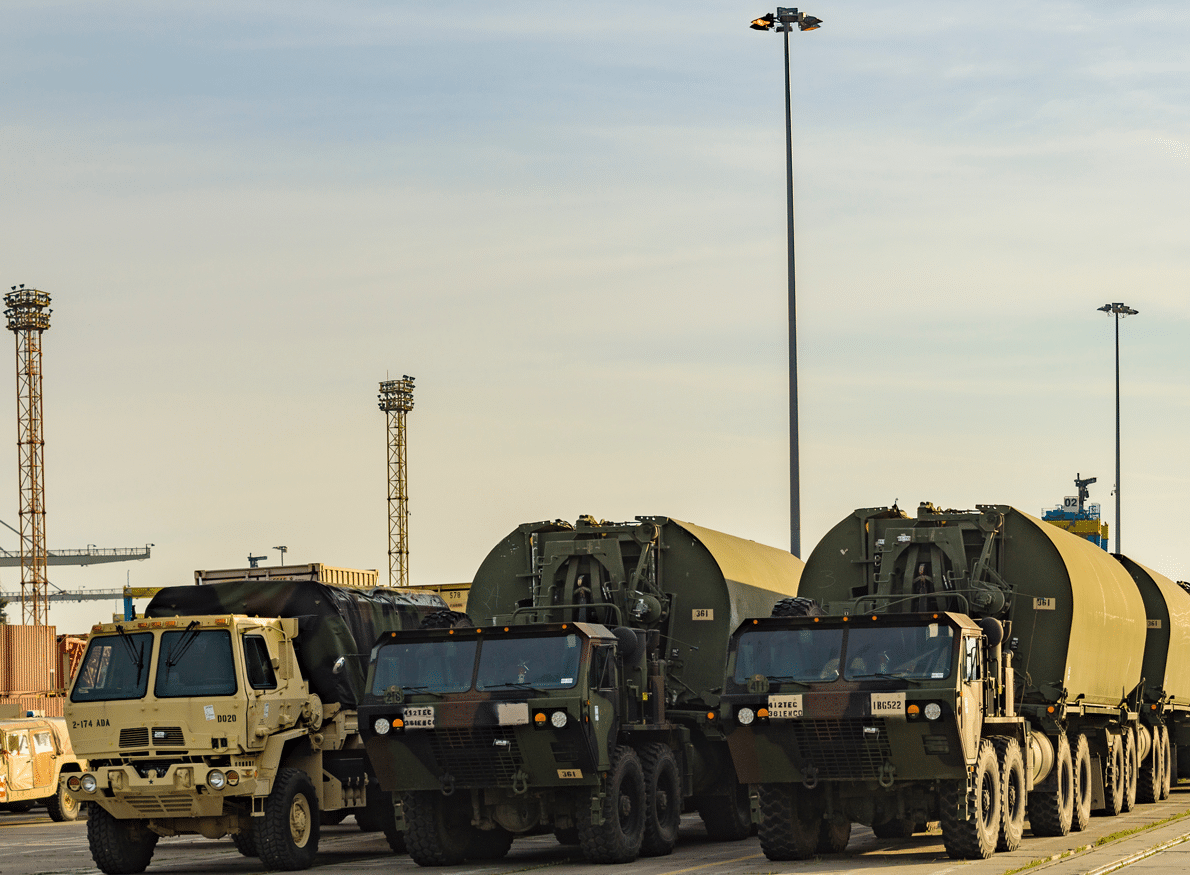 Military trucks lined up with large cargo containers, possibly at a port, under a hazy sky.