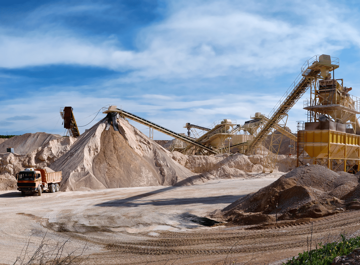 A quarry with conveyor belts, piles of sand, and a dump truck under a blue sky with clouds.
