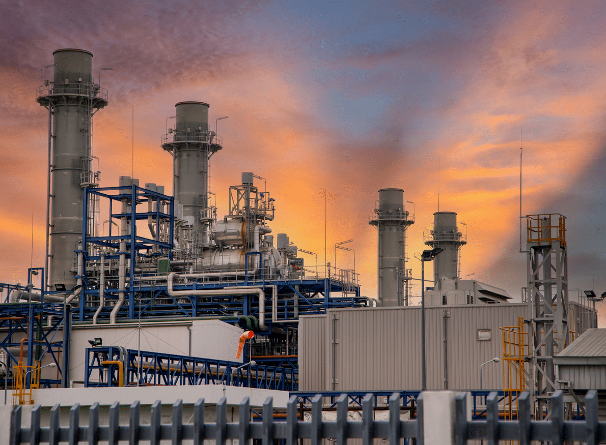 A power plant with tall stacks and intricate industrial machinery stands against a dramatic sunset sky with orange and purple clouds.