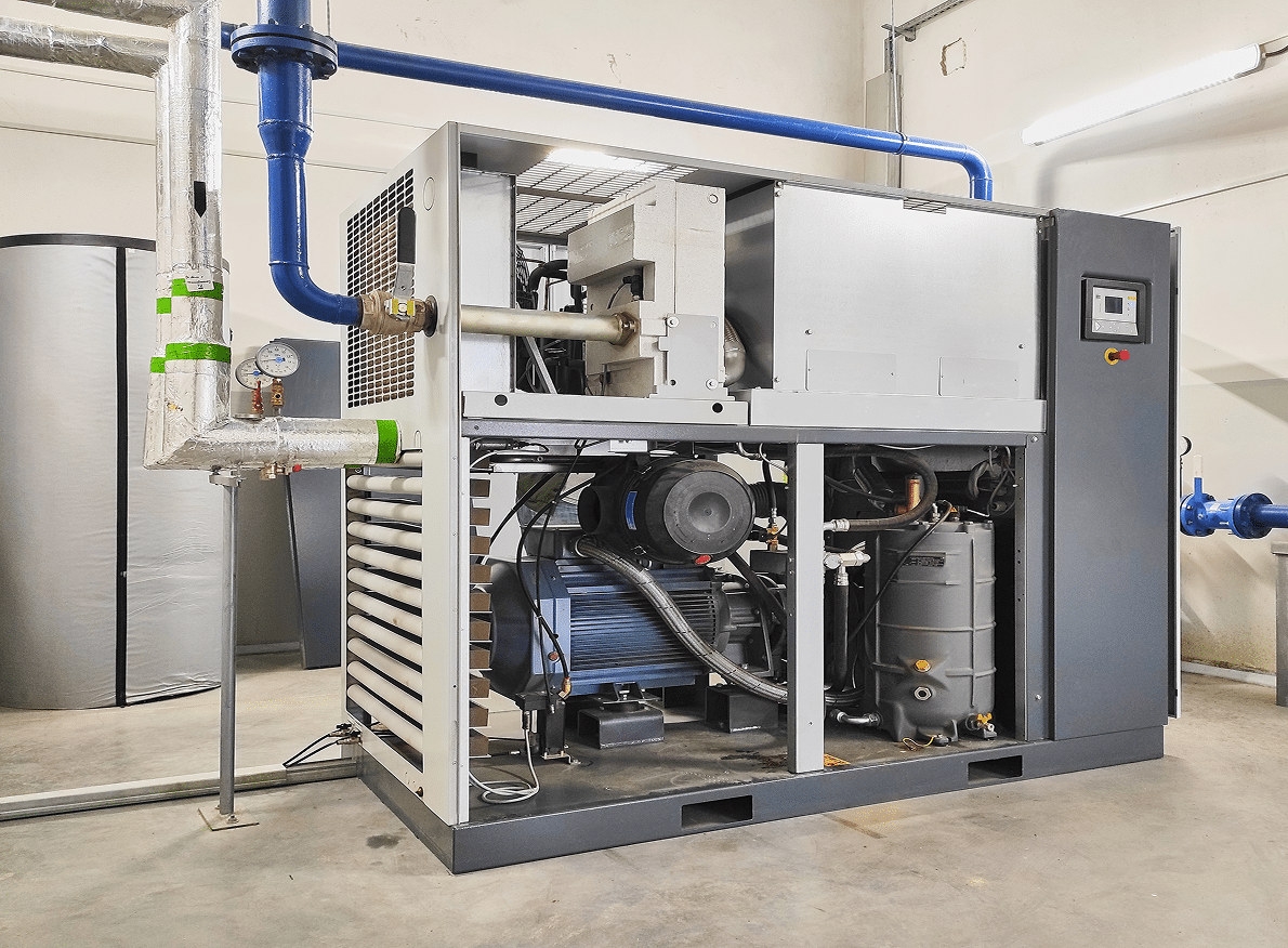 Industrial air compressor showing exposed mechanical parts and pipes in a bright, clean room with concrete floor and white walls.