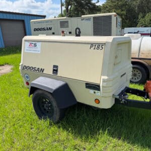 A beige Doosan P185 air compressor trailer with black wheels is on grass by a building; another compressor and white tank in background.