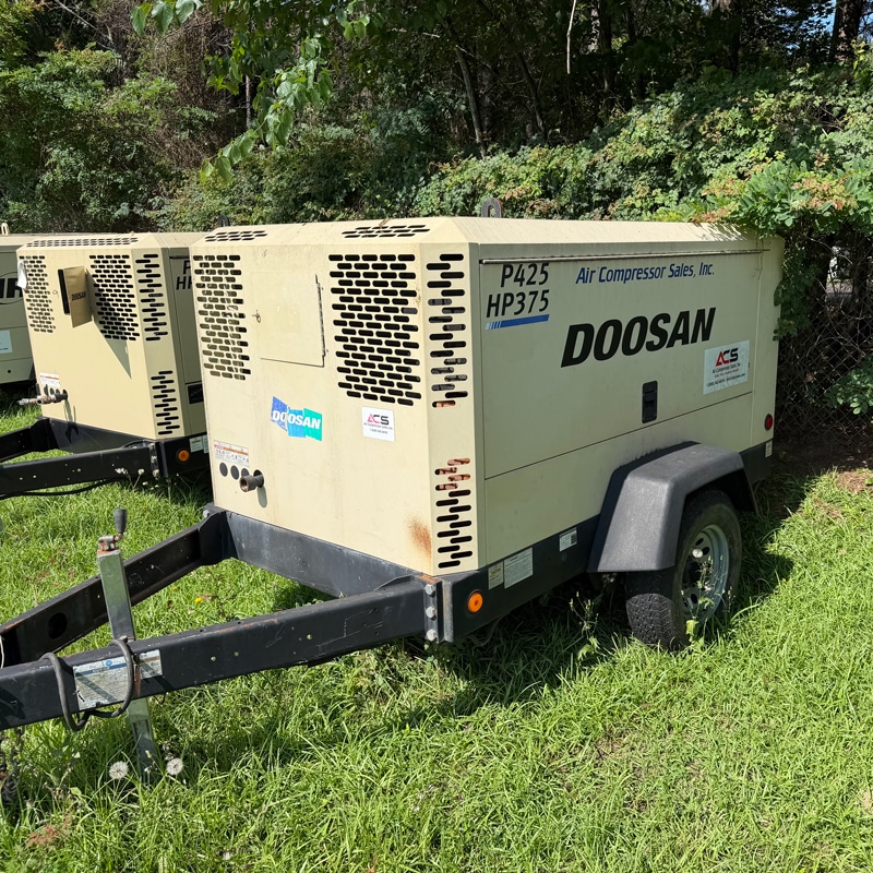 Two beige Doosan P425 HP375 air compressors on trailers parked on grass, trees visible in the background.