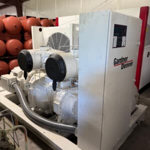 A large industrial air compressor labeled Gardner Denver with control panel, cooling fan, and pipes sits indoors with orange tanks behind.