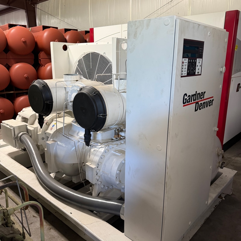 A large industrial air compressor labeled Gardner Denver with control panel, cooling fan, and pipes sits indoors with orange tanks behind.