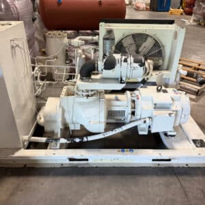 White industrial refrigeration compressor with pipes, fan, and motor on a warehouse floor among other equipment and supplies.