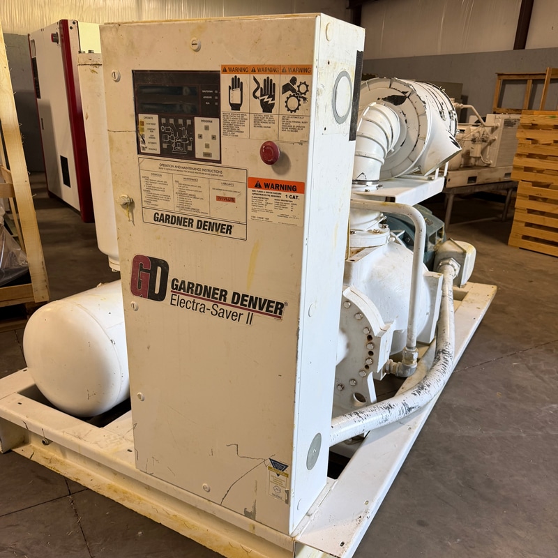 A white industrial air compressor labeled Gardner Denver Electra-Saver II sits on a warehouse floor with visible labels and control panel.