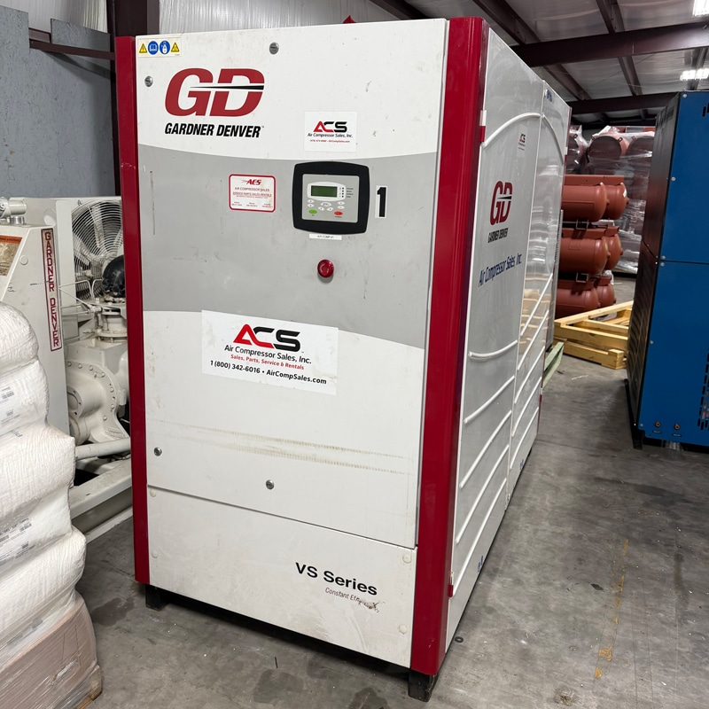 A large Gardner Denver VS Series industrial air compressor with digital controls and ACS branding sits in a warehouse amid equipment and pipes.
