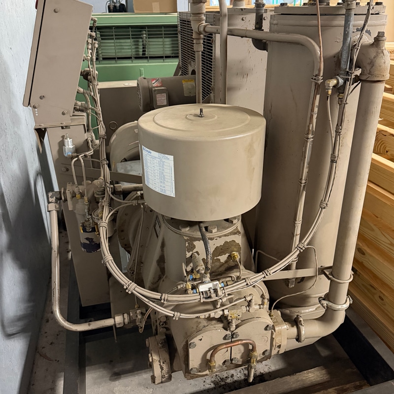 A large industrial air compressor with pipes, gauges, and control panel stands on concrete near a wooden crate against a gray wall.