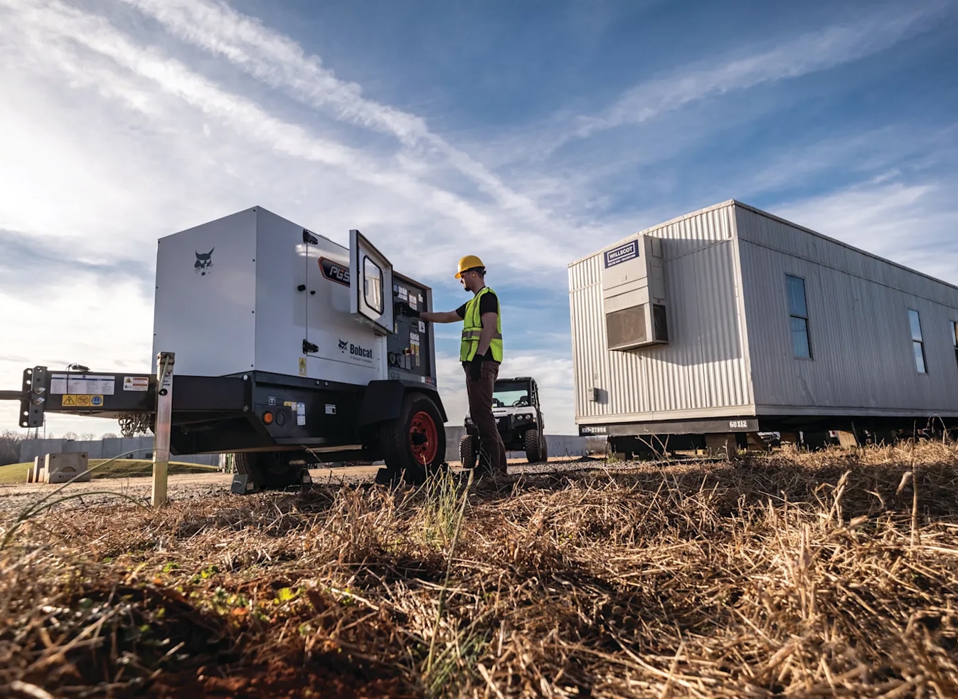 Construction worker wearing a yellow hard hat and vest runs a generator on dry grass next to a trailer under partly cloudy skies.